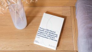 An overhead shot of a white report lying on a light wooden table. The report's title reads, "Thrive LDN Mental Health and Wellbeing Support for Displaced and Crisis-Affected Communities: A Review". To the left of the report is a glass vase with dried twigs, and to the right is the edge of a grey fabric cushion.